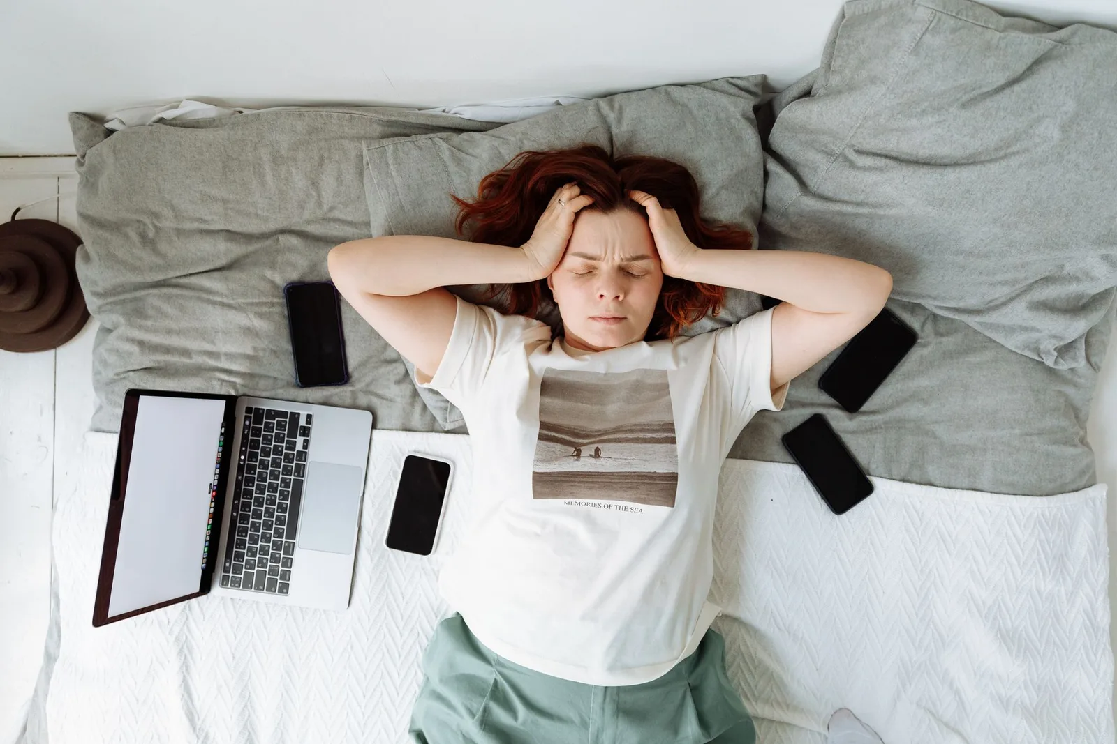 A stressed creator lying in bed with eyes shut, surrounded by phones and a laptop